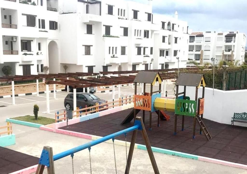 Un terrain de jeux avec des balançoires et des toboggans, entouré d’un parking et bordé d’une rangée d’immeubles d’habitation blancs.