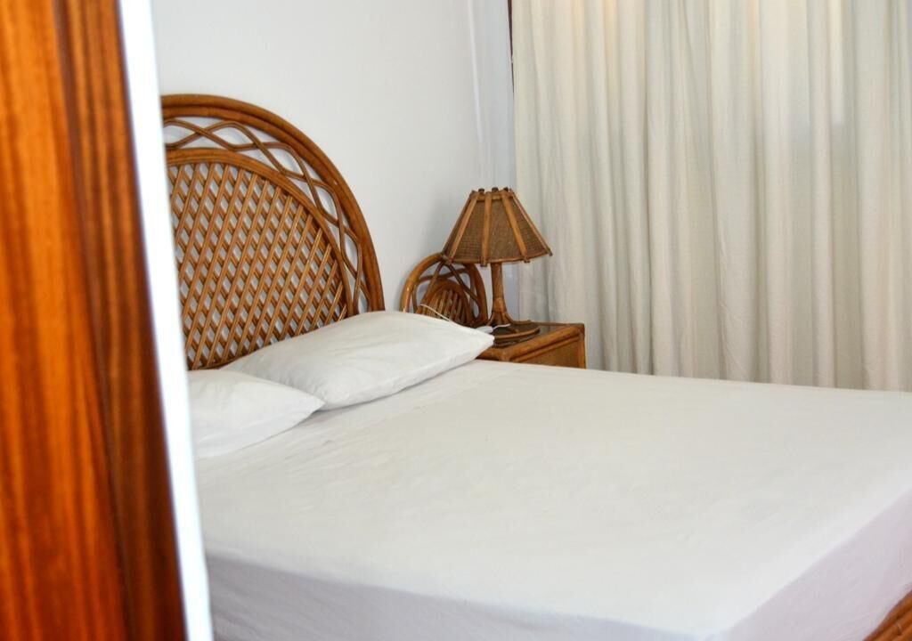 A neatly made bed with white linens, a wicker headboard, and a bedside lamp on a wooden nightstand.