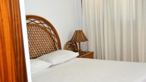 Een net opgemaakt bed met witte lakens, een rieten hoofdbord en een bedlampje op een houten nachtkastje.