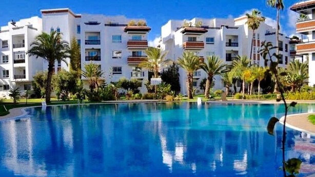Una piscina rodeada de edificios de varios pisos con balcones, palmeras y un cielo azul despejado.
