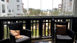 Un balcón con sillas de mimbre y una pequeña mesa, con vistas a una zona residencial con palmeras y edificios.