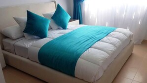 Ein Bett mit weißer Bettwäsche und Türkis-Akzenten, ein Kopfteil und ein Fenster mit Vorhängen.