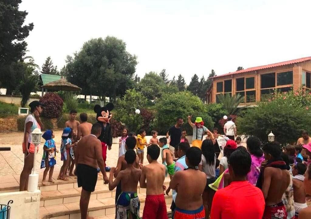 A group of people gathered outdoors, some wearing swimwear, in a courtyard with steps leading up to a building with large windows.