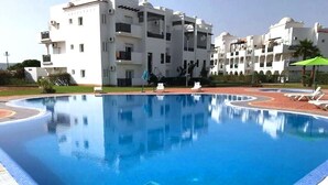 A swimming pool surrounded by white buildings with balconies, a clear blue sky, and a few lounge chairs.