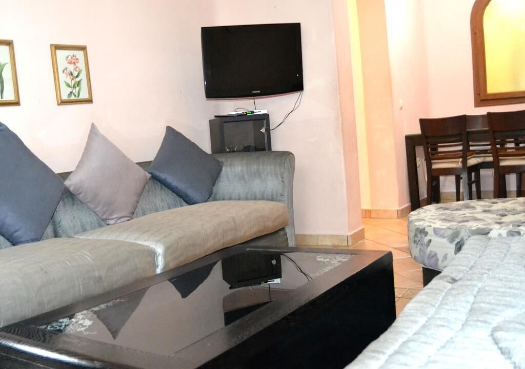 Una sala de estar con sofá, televisor de pantalla plana montado en la pared, una mesita y una cama con colcha estampada.