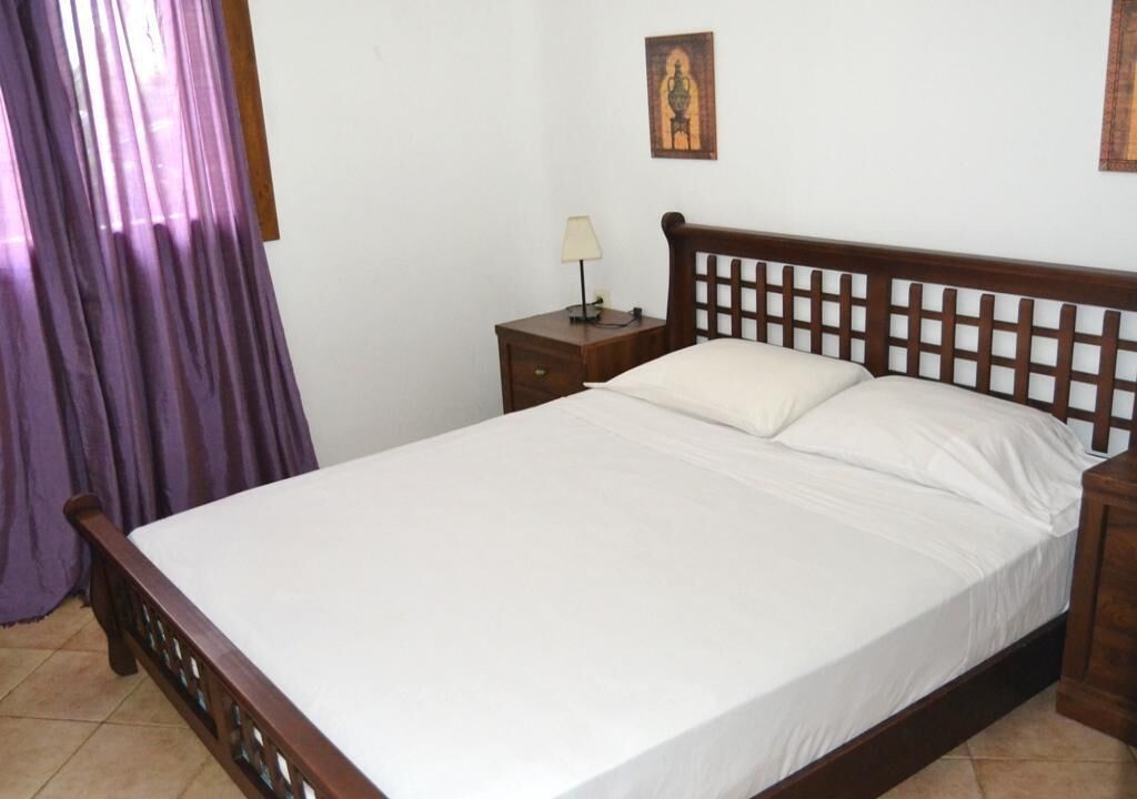 Una cama bien hecha con cabecera de madera, una mesita de noche con lámpara y un cuadro colgado en la pared.