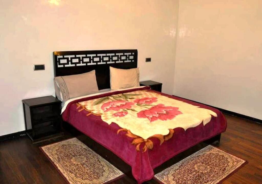 Un lit bien fait, recouvert d’une couvre-lit à motifs floraux, deux tables de chevet et deux tapis sur un plancher en bois.