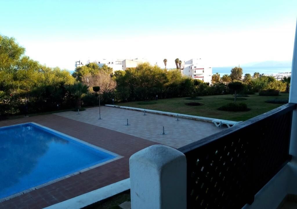 A swimming pool with a tiled deck, surrounded by greenery and a view of buildings in the distance.