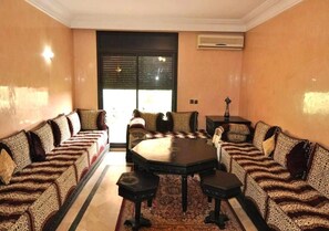 A room with a central round table surrounded by sofas, a patterned rug, and a large window with blinds.