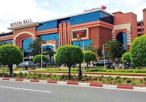 Un centro comercial moderno con un llamativo letrero de 'Menara Mall', un anuncio de 'New Burger' y jardines bien cuidados con árboles podados y una carretera despejada al frente.