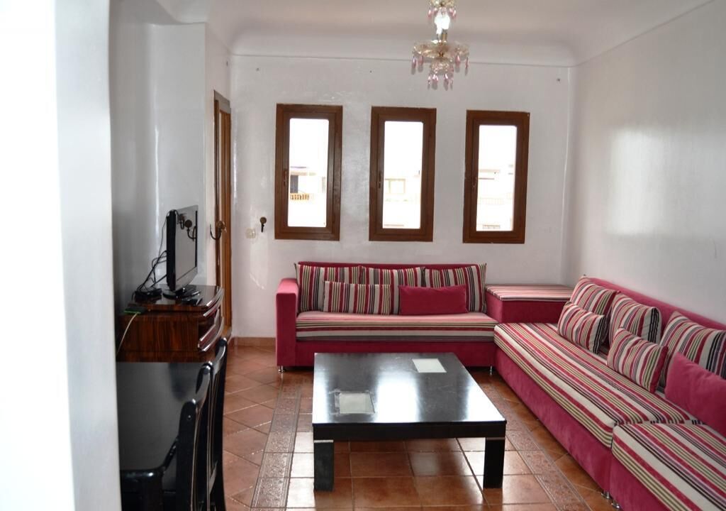Ein Wohnzimmer mit einer Couch, einem Couchtisch und einem Fernseher. Drei Fenster und eine Deckenleuchte sind vorhanden.