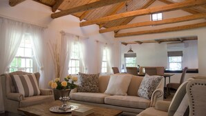 A cozy living room with a wooden ceiling, a sofa set, a coffee table with a vase of flowers, and a dining area in the background. - La Paris Estate (Franschhoek)