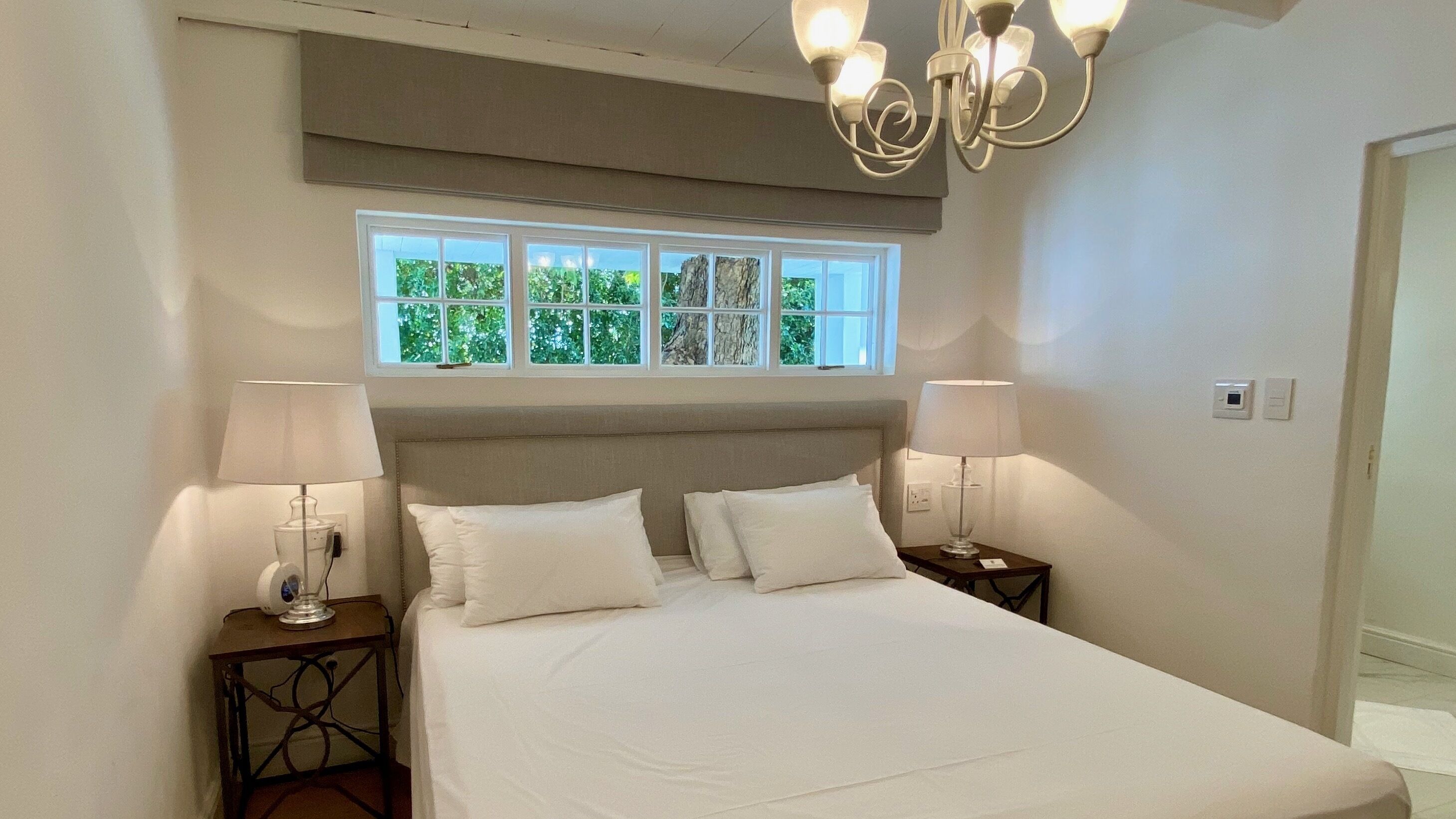 A bedroom with a large bed, two bedside tables with lamps, a window with a view of trees, and a chandelier.
