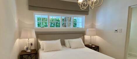 A bedroom with a large bed, two bedside tables with lamps, a window with a view of trees, and a chandelier.