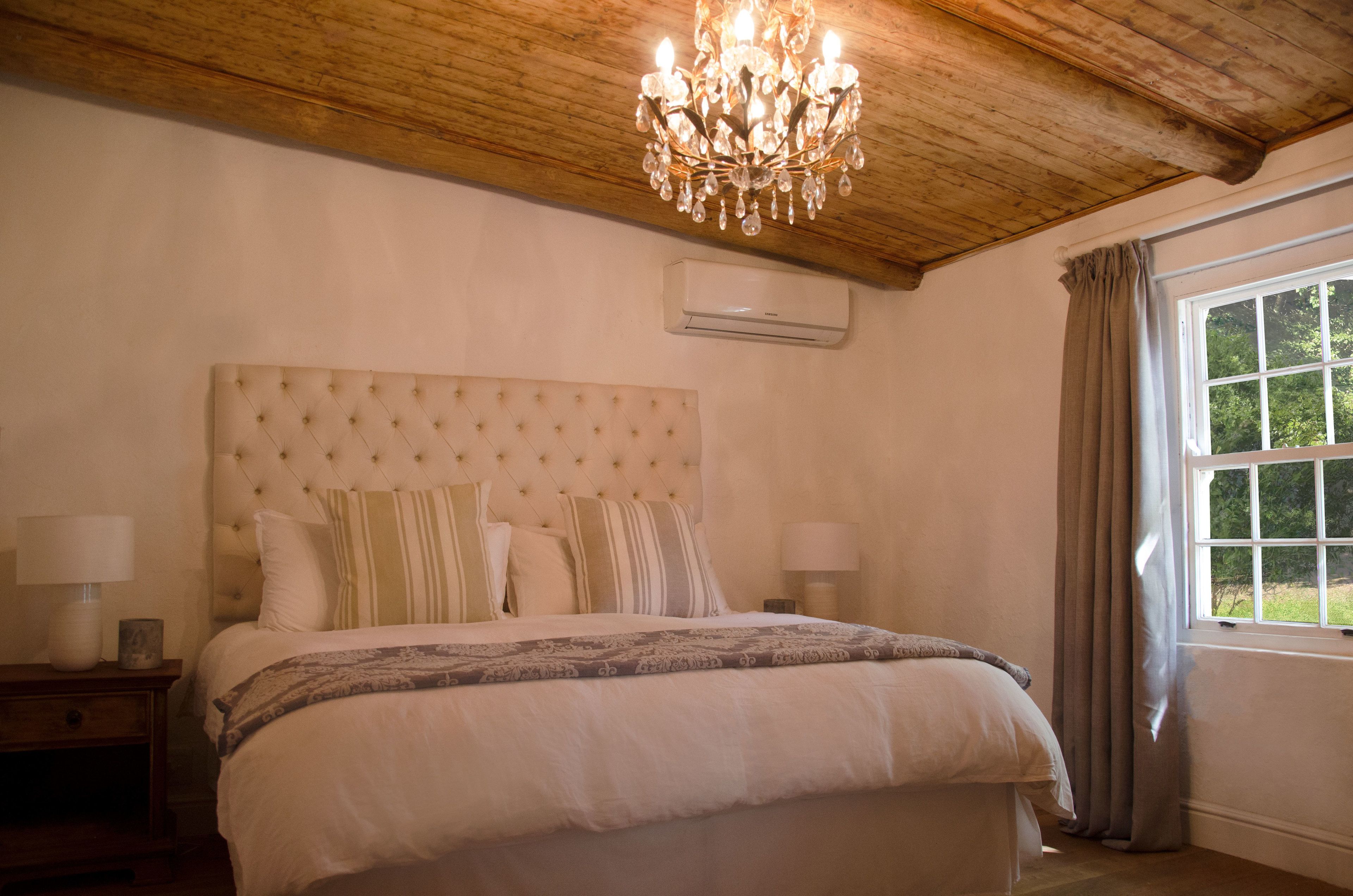 A bedroom with a tufted headboard, a chandelier, a window with curtains, and an air conditioning unit.