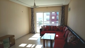 A living room with a red sofa, a glass coffee table, and a cabinet. There is a large window with curtains and a view of buildings outside. - Ocean Atlantic Residence 3050 (Asilah)