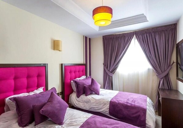 Une chambre d’hôtel avec deux lits, chacun doté d’une tête de lit capitonnée, de draps violets et d’une grande fenêtre ornée de rideaux violets.
