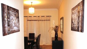 Uma sala de jantar com uma mesa, cadeiras, um vaso com plantas secas e duas pinturas abstratas nas paredes.