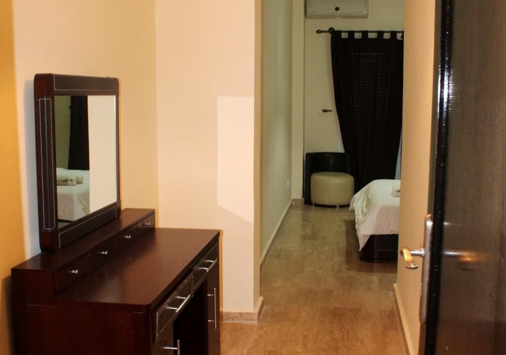 A hotel room with a wooden dresser, a bed with a white bedspread, a black curtain, and a small round ottoman.