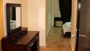 Hotellihuone, jossa on puinen lipasto, valkoisella peitteellä varustettu sänky, musta verho ja pieni pyöreä rahi.