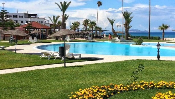 A pool area with lounge chairs, palm trees, and a building in the background. - Residence Marina Beach Mdiq 3402 (Mdiq)