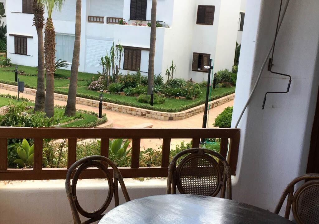 Un balcón con sillones de mimbre y una mesa, con vistas a un jardín cuidado que incluye palmeras y un edificio blanco al fondo.