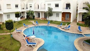 Une piscine entourée de chaises longues bleues et de parasols, le tout bordé d’un bâtiment blanc doté de fenêtres en arc de cercle et de balcons.