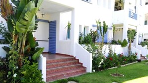 Un edificio blanco con contraventanas azules, una escalera que lleva a un porche, y un jardín bien cuidado con diversas plantas y flores.