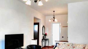 Una sala de estar con una televisión de pantalla plana, un sillón negro, un sofá floral, una mesita pequeña y un espejo decorativo.