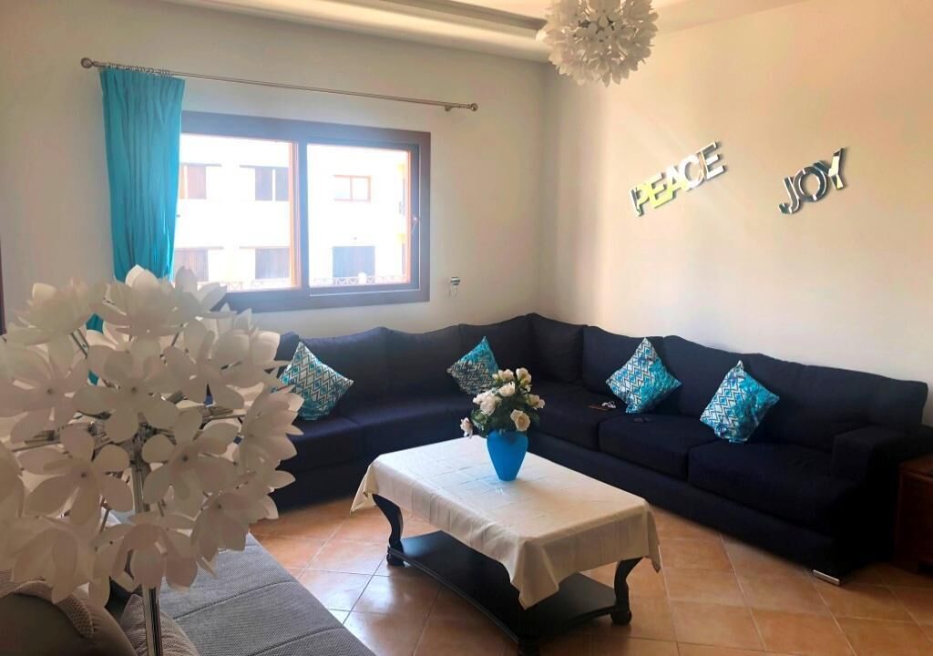A living room with a dark blue sofa, a white table with a vase of flowers, and a window with blue curtains.