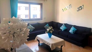 A living room with a dark blue sofa, a white table with a vase of flowers, and a window with blue curtains. - Lilac's Garden Residence 2947 (Cabo Negro)