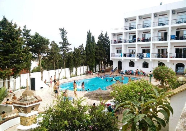 A hotel with a swimming pool surrounded by greenery, a white building with multiple balconies, and a stone structure near the pool.