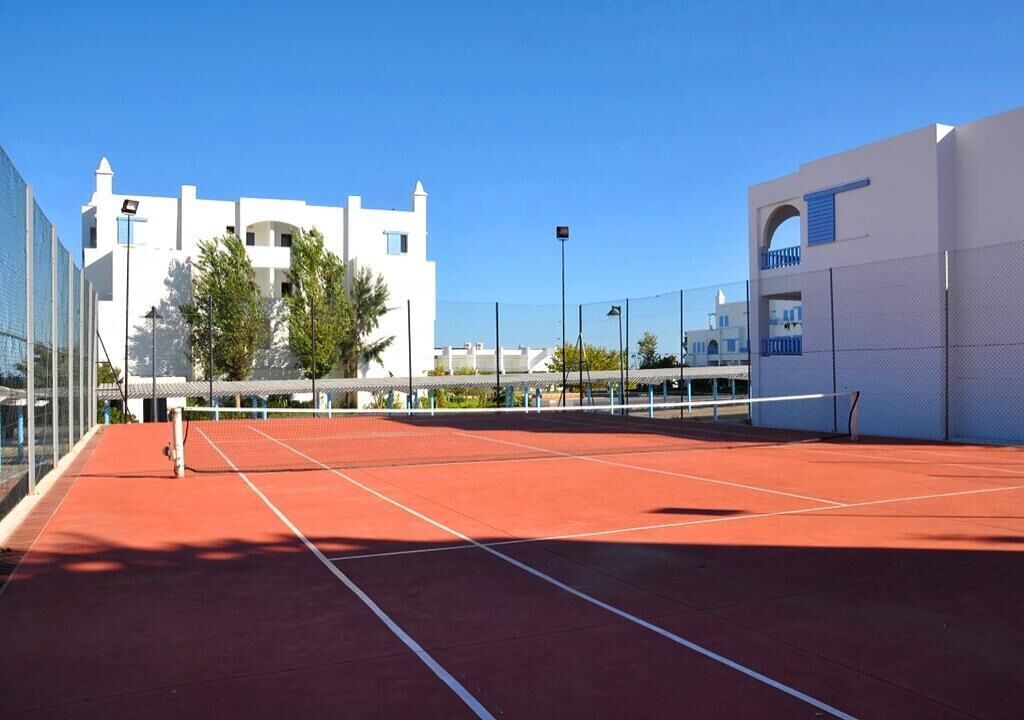 An outdoor tennis court with a red surface, surrounded by modern buildings and a clear blue sky.