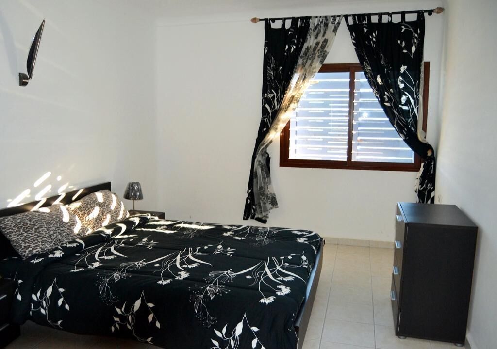 A bedroom with a bed, a nightstand, a dresser, a window with curtains, and a wall-mounted lamp.
