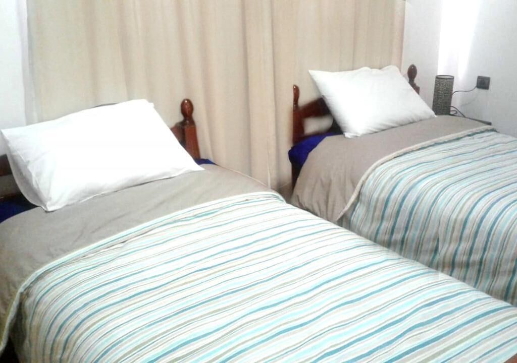 Two single beds with wooden frames and white pillows, a beige curtain, and a small table with a lamp.