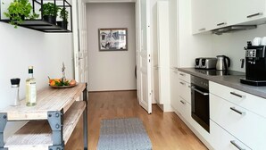 Ett modernt kök med vita skåp, en inbyggd ugn och en kaffemaskin. Det finns ett träbord med en flaska och en skål med frukt. En svart metallhylla rymmer växter, och en inramad tavla hänger på väggen.