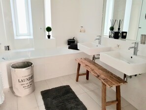 Moderni kylpyhuone, jossa on suuri kylpyamme, kaksi pesuallasta, puinen penkki, ruukussa oleva kasvi ja ämpäri, jossa on merkintä ”PYYKKI”.