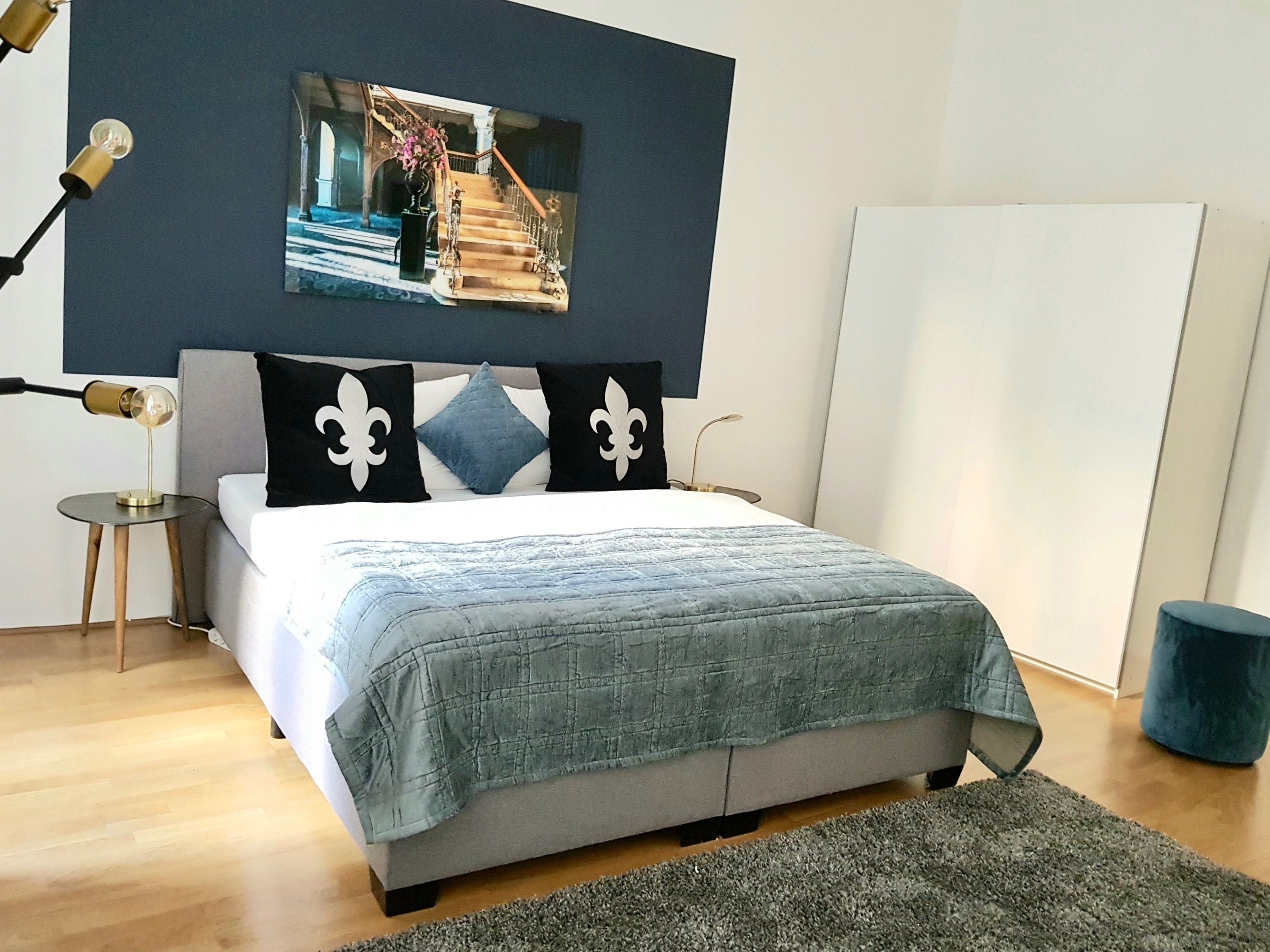 Ein modernes Schlafzimmer mit einem Bett, einem kleinen Nachttisch, einer Lampe, einem Bild an der Wand und einem blauen Sessel.