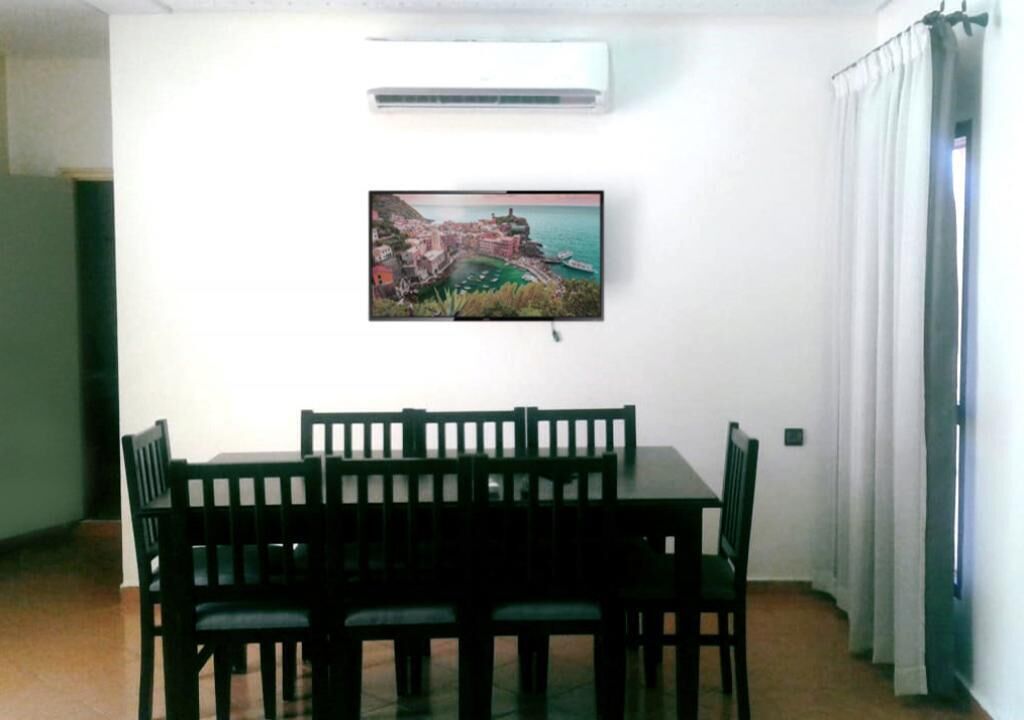 A dining area with a table and chairs, an air conditioning unit, and a painting of a coastal town on the wall.