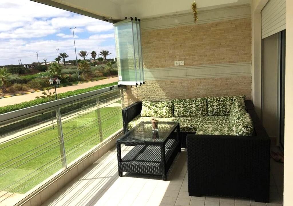A balcony with a sofa and a glass table, overlooking a green landscape with palm trees.