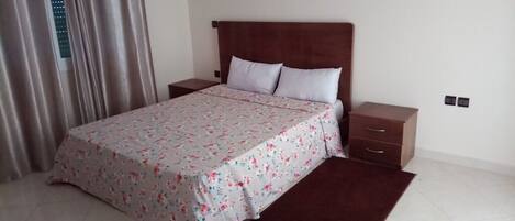 A bedroom with a bed, two nightstands, a wooden headboard, a floral bedspread, a brown rug, and a window with curtains.