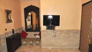 A living room with a TV mounted on a brick wall, a dark wooden cabinet, and a red pillow on the floor. - Residence Barrone Marrakech 3384 (Marrakech)