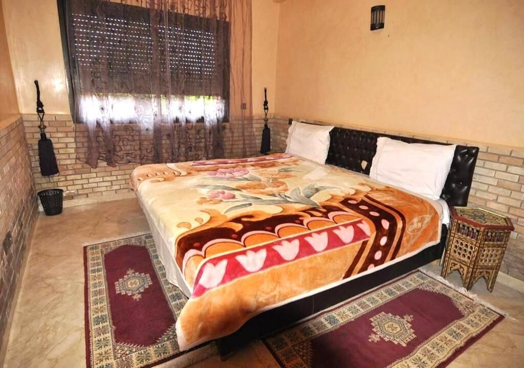 Une chambre à coucher avec un grand lit, une couvre-lit à motifs, une fenêtre avec des rideaux, une petite table de chevet et un tapis décoratif.