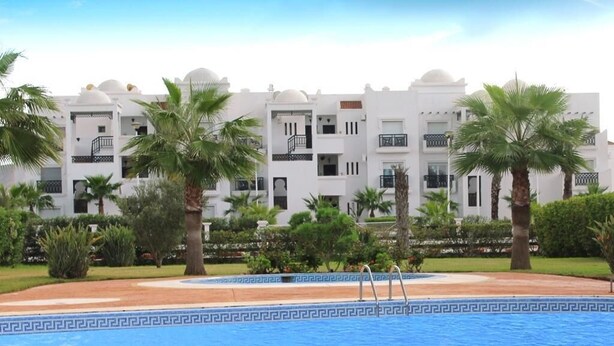 A swimming pool with a geometric tile pattern, surrounded by palm trees and a white building with balconies. - The Greens Residence 3633 (SAIDIA)