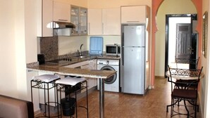 Cocina compacta con microondas, refrigerador y lavadora. Hay dos taburetes y una mesa pequeña.
