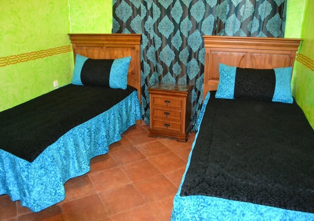 Una habitación con dos camas, cada una con cabecero de madera y colcha azul. Entre las camas hay una mesita de noche de madera. Las paredes están pintadas de verde y el suelo está cubierto de baldosas de terracota.
