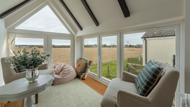 A cozy room with a high ceiling, large windows, a white table with a vase, a beige sofa, and a patterned cushion. - First & Last, Trimingham, Norfolk (Trimingham)