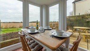 A dining table set for four with white plates, bowls, and cutlery, positioned in a room with large windows overlooking a garden. - First & Last, Trimingham, Norfolk (Trimingham)