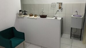 A kitchen with a white countertop, a sink, and a small refrigerator. There are various kitchen items on the counter, including a kettle, a basket, and a bowl. A green chair is visible in the foreground. - Espaço da Caraputinga (São Paulo)
