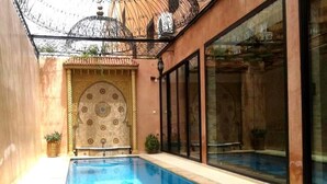 Ein Schwimmbecken mit befestigtem Beckenrand, eine dekorative Wand mit kreisförmigem Muster und eine Glastür zu einem überdachten Bereich.